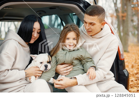Family resting after day spending outdoor in autumn park. Father, mother, their daughter and dog sitting inside car trunk, smiling. Family wearing beige sportive costumes. Family resting after day spending outdoor in autumn park. Father, mother, their daughter and dog sitting inside car trunk, smiling. Family wearing beige sportive costumes. 128845051