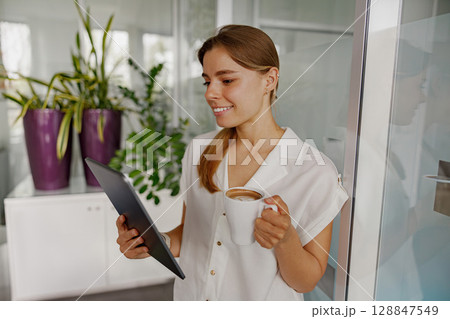 A Young Woman Delightfully Enjoying Her Coffee While Using a Tablet in a Modern, Stylish Space 128847549