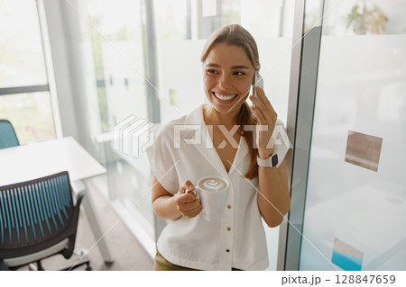 A smiling woman in a modern office is talking on the phone while enjoying her coffee beverage A smiling woman in a modern office is talking on the phone while enjoying her coffee beverage 128847659