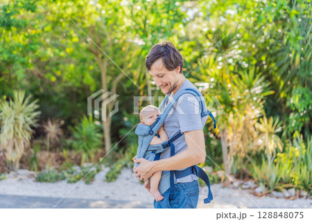 Young father walks in nature with his baby son nestled close in a parent-facing ergonomic baby carrier. Bonding, warmth, and modern parenting concept in a peaceful outdoor setting 128848075