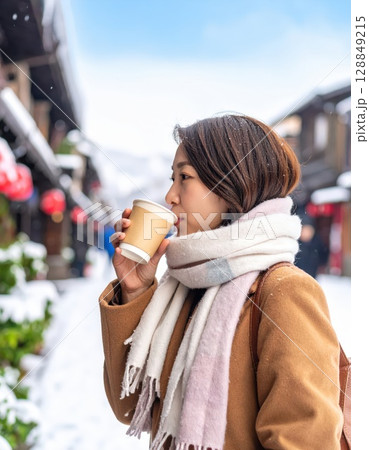 《AI画像》雪の積もる繁華街でホットドリンクを飲むコートを着た若い女性 128849215