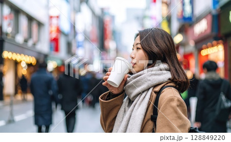 《AI画像》雪の積もる繁華街でホットドリンクを飲むコートを着た若い女性 128849220