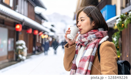 《AI画像》雪の積もる繁華街でホットドリンクを飲むコートを着た若い女性 128849226