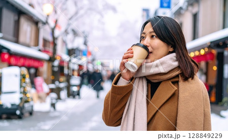 《AI画像》雪の積もる繁華街でホットドリンクを飲むコートを着た若い女性 128849229