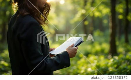 Close-up of young businesswoman writing in notebook while standing in green forest Close-up of young businesswoman writing in notebook while standing in green forest 128850572