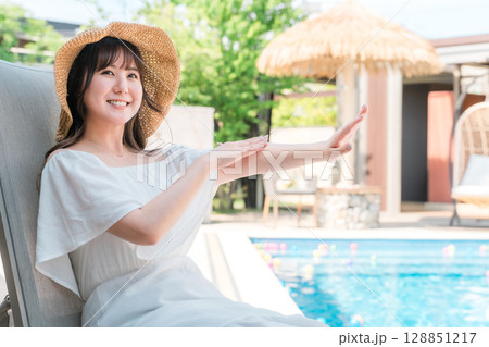 プールサイドのリクライニングチェアに座り日焼け止めクリームを腕に塗る若いアジア人女性 プールサイドのリクライニングチェアに座り日焼け止めクリームを腕に塗る若いアジア人女性 128851217