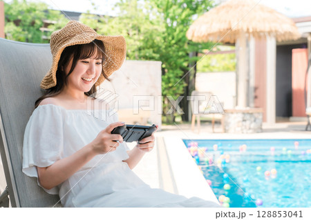 プールサイドでリクライニングチェアに座りゲームをする若いアジア人女性 128853041