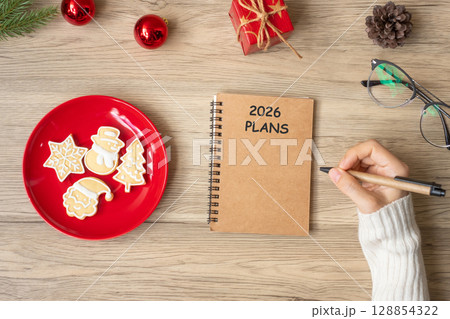 Woman hand writing 2026 PLANS on notebook with Christmas cookies on table. Xmas, Happy New Year, Goals, Resolution, To do list, and Strategy concept 128854322