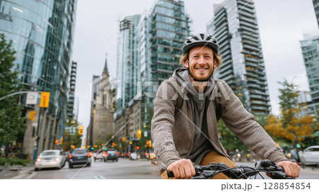 Smiling young man riding bicycle in modern city with glass buildings and traffic around. Urban lifestyle and eco transport concept. 128854854