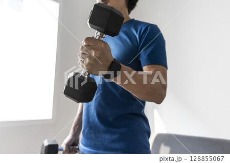 handsome man doing dumbbell workout at home, working on arms strength at living room interior. 128855067