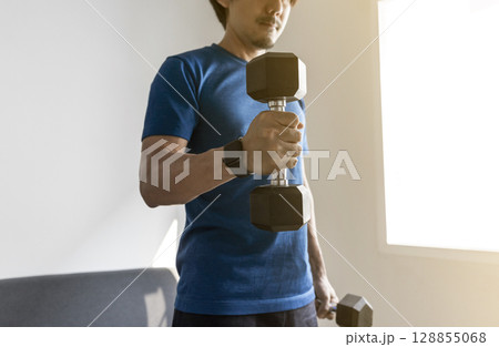handsome man doing dumbbell workout at home, working on arms strength at living room interior. 128855068