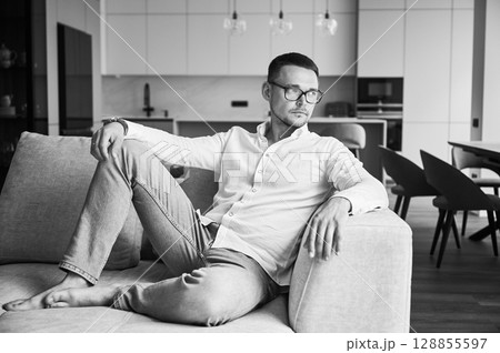 Charming man relaxing on couch in his apartment. Handsome young male thoughtfully looking away. Man in casual clothes lying on sofa on background of modern kitchen. Black and white image. Charming man relaxing on couch in his apartment. Handsome young male thoughtfully looking away. Man in casual clothes lying on sofa on background of modern kitchen. Black and white image. 128855597