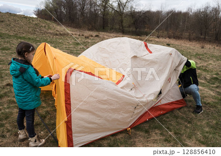Children explore outdoor adventures while setting up a colorful tent in a tranquil field under a cloudy sky during early springtime Children explore outdoor adventures while setting up a colorful tent in a tranquil field under a cloudy sky during early springtime 128856410