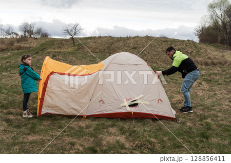 Setting up a cozy tent on a grassy hillside under a cloudy sky, with a child eagerly helping while enjoying outdoor adventure together Setting up a cozy tent on a grassy hillside under a cloudy sky, with a child eagerly helping while enjoying outdoor adventure together 128856411