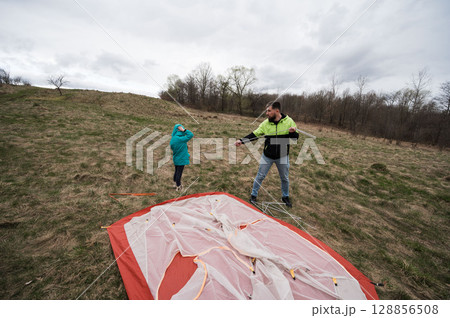 Father and child share a bonding moment while setting up a tent in a scenic meadow under an overcast sky during early spring, embracing nature and adventure 128856508