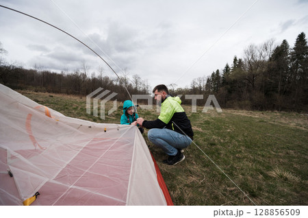 A father and daughter are enjoying quality time together as they work to set up a tent beneath a cloudy sky in a serene park on a chilly day, creating lasting memories in the great outdoors 128856509