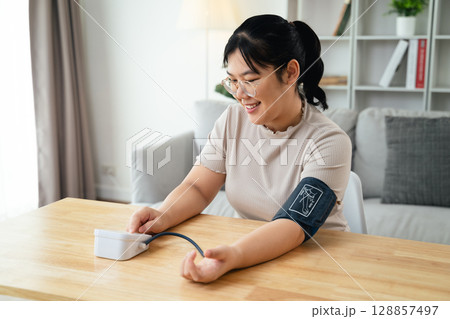 An Asian woman uses a portable blood pressure monitor in her living room. Health awareness concept. An Asian woman uses a portable blood pressure monitor in her living room. Health awareness concept. 128857497