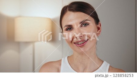 Portrait of a young Caucasian woman wearing braces, smiling warmly in home interior. Represents dental care and orthodontics. Bright, inviting atmosphere with soft lighting in background. Portrait of a young Caucasian woman wearing braces, smiling warmly in home interior. Represents dental care and orthodontics. Bright, inviting atmosphere with soft lighting in background. 128858253