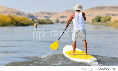 Young male paddleboarding on calm river surrounded by scenic hills, rear view 128859119