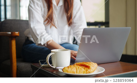 front view of woman using laptop computer, working from home in the morning with hot coffee, croissant bakery and mobile phone on the table. anonymous face front view of woman using laptop computer, working from home in the morning with hot coffee, croissant bakery and mobile phone on the table. anonymous face 128859364