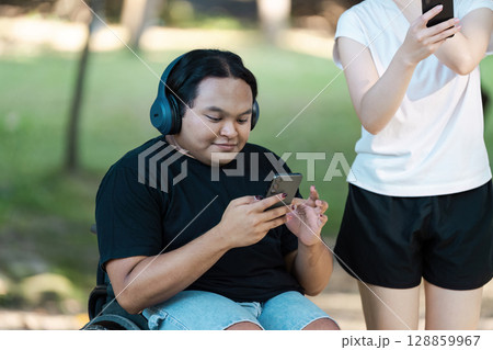 Young Man in Wheelchair Enjoying Music on Headphones While Using Smartphone Outdoors with Friend in Casual Summer Attire 128859967