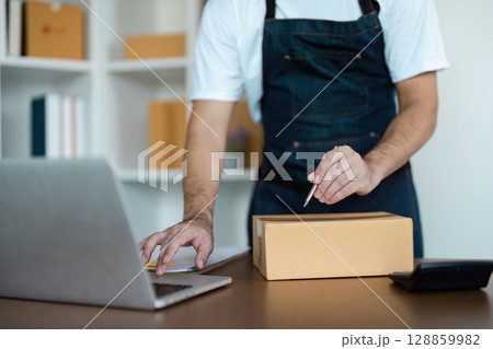 E-commerce Business Owner Preparing Package for Shipping with Laptop and Cardboard Box in Modern Office Workspace 128859982