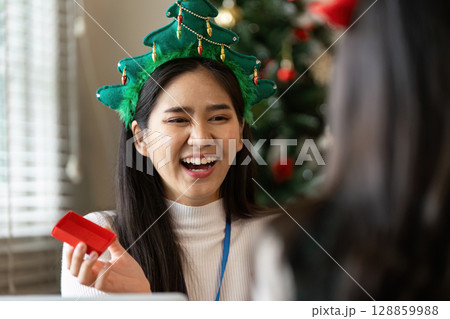 Festive Office Celebration with Smiling Employee in Christmas Tree Hat Exchanging Gifts in a Modern Workplace Setting 128859988