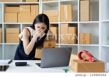 Stressed Young Woman Managing Online Business with Laptop and Cardboard Boxes in Home Office Stressed Young Woman Managing Online Business with Laptop and Cardboard Boxes in Home Office 128860268