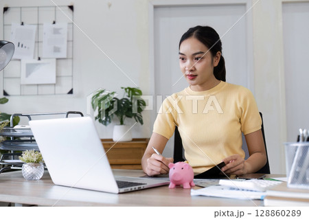 Young Professional Woman Engaged in Financial Planning with Laptop and Piggy Bank for Savings in Modern Office Setting 128860289
