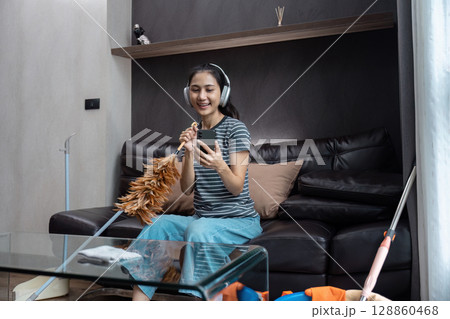 Smiling Housekeeper Enjoying Music While Cleaning Living Room with Headphones and Duster in Modern Home Environment 128860468