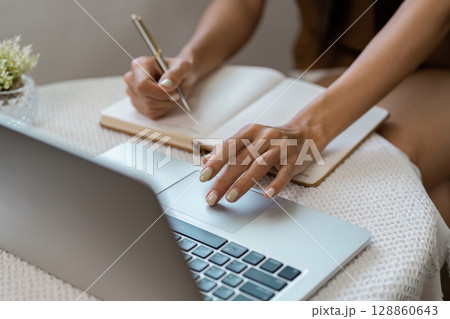 Woman Writing in Notebook While Using Laptop at Home Office for Remote Work or Study in Cozy Environment 128860643
