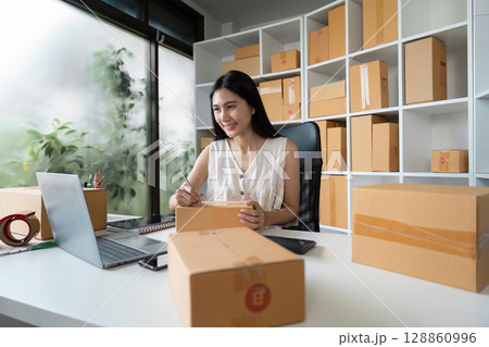 Young Entrepreneur Working on Online Orders in a Modern Home Office with Shipping Boxes and Laptop 128860996