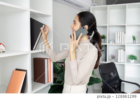 Businesswoman Organizing Bookshelf While Talking on Smartphone in Modern Office Setting 128861299