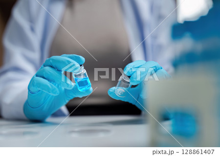 Scientist in Laboratory Holding Vials with Blue Liquid, Wearing Protective Gloves and Lab Coat, Conducting Research Scientist in Laboratory Holding Vials with Blue Liquid, Wearing Protective Gloves and Lab Coat, Conducting Research 128861407