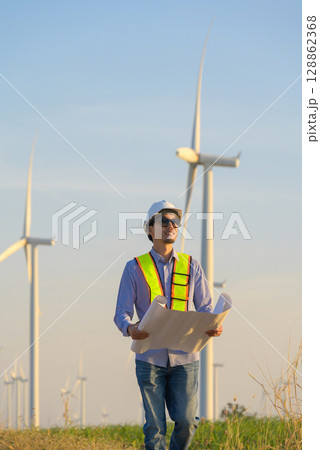 Japanes engineer in white helmet holding blueprint in wind farm for assessing or monitoring wind turbine equipment Japanes engineer in white helmet holding blueprint in wind farm for assessing or monitoring wind turbine equipment 128862368