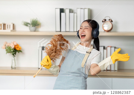 Happy Cleaning. Young woman smiling while holding a feather duster and headphones. Happy Cleaning. Young woman smiling while holding a feather duster and headphones. 128862387
