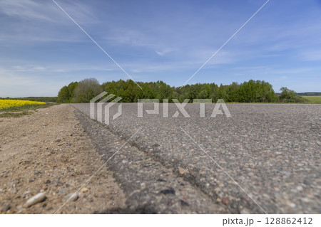 an asphalt road in a field on one side grows flowering rapeseed, and on the other side grows green grass against a blue sky with clouds 128862412