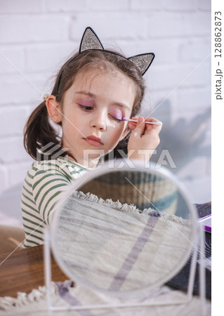 Young girl with cat ear headband applies bright pink eyeshadow with a cotton swab, deeply concentrated and enjoying her makeup play Young girl with cat ear headband applies bright pink eyeshadow with a cotton swab, deeply concentrated and enjoying her makeup play 128862873