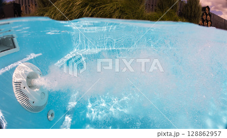 Underwater view of a jet stream creating intense bubbles in a backyard pool with decorative greenery and wooden fencing in the background. 128862957
