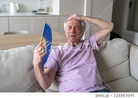 No cooling system, elderly man struggling without air conditioning, resting on couch using fan No cooling system, elderly man struggling without air conditioning, resting on couch using fan 128863045