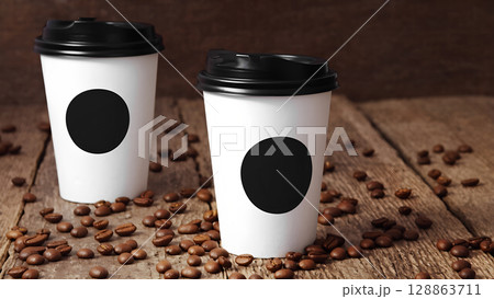 Two white coffee cups with black lids are placed on a wooden table, which is covered with coffee beans. The cups are filled with a white liquid, possibly coffee 128863711