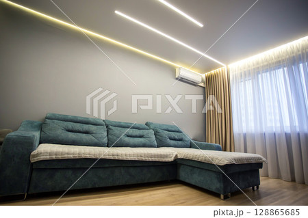 Interior in the hall: blue large sofa against the background of a window with brown curtains and wooden laminate. Modern interior room hall. Copy space for text Interior in the hall: blue large sofa against the background of a window with brown curtains and wooden laminate. Modern interior room hall. Copy space for text 128865685