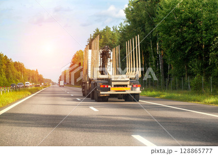 An empty timber truck with a hydraulic manipulator goes into the forest for timber along the highway in the summer against the backdrop of the sun. Forest industry concept. Copy space for text An empty timber truck with a hydraulic manipulator goes into the forest for timber along the highway in the summer against the backdrop of the sun. Forest industry concept. Copy space for text 128865777