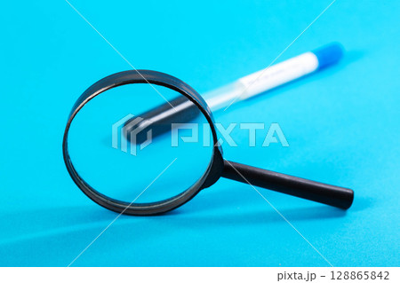 A magnifying glass against the background of a sterile test tube with an analysis for vitamins and a blue background. Analysis for a complex of vitamins, micro and macroelements, close-up A magnifying glass against the background of a sterile test tube with an analysis for vitamins and a blue background. Analysis for a complex of vitamins, micro and macroelements, close-up 128865842