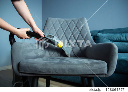 The hands of a girl with a washing vacuum cleaner clean and wash the gray upholstery of a soft chair from dirt. Cleaning services, business, close-up. Copy space for text 128865875