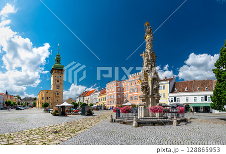 Panoramic view of the main square in Retz, Austria, with Holy Trinity Column, colorful buildings, town hall tower, blooming flowers and warm summer atmosphere. 128865953