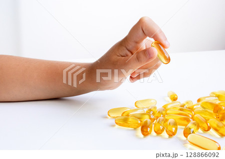 Child hand holding omega 3 capsule amidst scattered supplements on white background 128866092