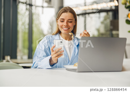 A Happy and Smiling Woman Enjoying Her Breakfast While Browsing on Her Smartphone 128866454