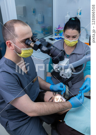 Dental professionals conduct a precise examination using a microscope while ensuring patient comfort and safety through proper protocols in a modern dental clinic environment. Dental professionals conduct a precise examination using a microscope while ensuring patient comfort and safety through proper protocols in a modern dental clinic environment. 128866508