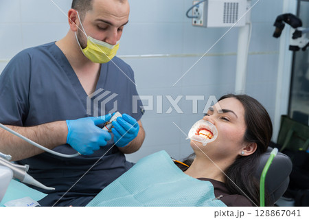 A Dental Procedure in Action: Dentist Preparing Tools While Patient Is Comfortable in the Chair, Showing Professionalism and Care in a Modern Clinic Setting. 128867041